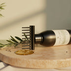 A black metal hair detangling comb with a wide, flexible bristle head, shown resting on a dark bottle of olive oil on a round marble board with a gold pool of oil and olive branches. Keshaura Anti Hairfall Hair Oil.