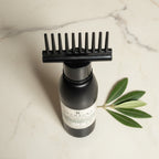 A black bottle with a black wide-tooth comb attachment resting on top, photographed on a light marble surface with a green leaf sprig beside it. The bottle label shows the brand Keshaura. Keshaura Anti Hairfall Hair Oil.