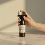 A hand holds a dark brown bottle with a black comb attachment against a warm beige background with soft shadows. Keshaura Anti Hairfall Hair Oil.